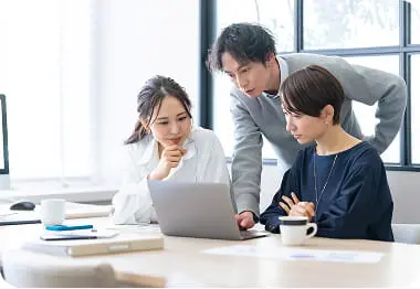 3人の男女がノートパソコンを見ながら話し合っているチームミーティングの様子