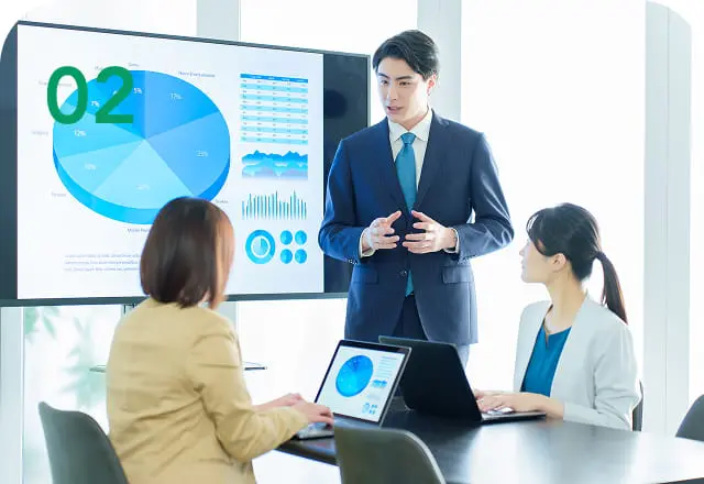 スーツ姿の男性がプレゼンテーションを行い、他の2人が資料を見ながら話を聞いている会議風景（画像