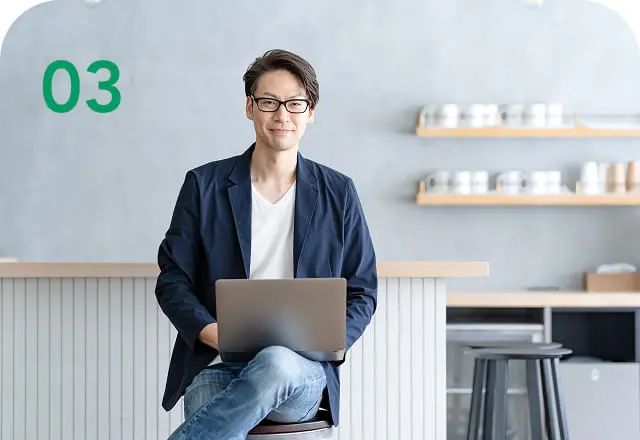 カフェ風の空間でノートパソコンを使いながら笑顔でカメラを見るカジュアルなビジネスマン