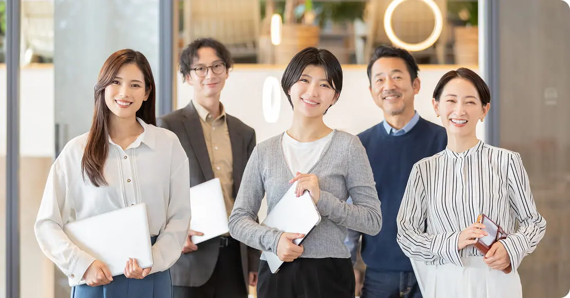 オフィスらしき空間でノートパソコンやタブレットを持った5人の男女が笑顔で並んでいる集合写真